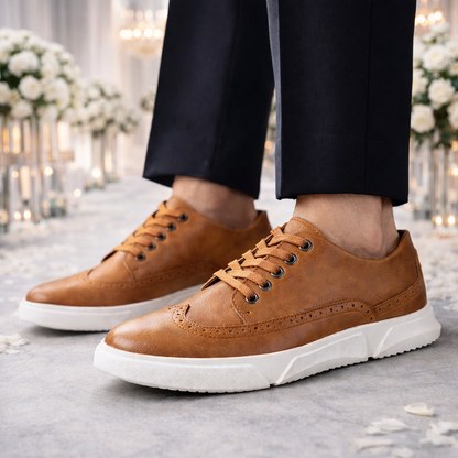 Men's tan leather Brogan lace-up sneakers with brogue detailing and white sole, paired with dark dress pants.