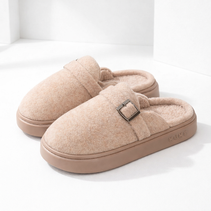 Cozy beige plush slip-on mule slippers with metal buckle detail and thick rubber sole, labeled HOME
